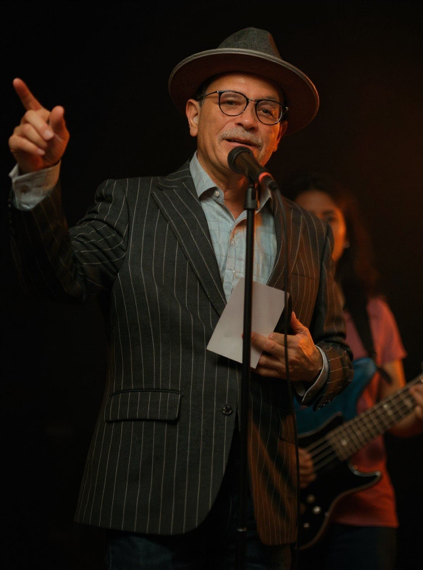 Daryl Sweeney on stage at a First Fridays concert, hat on, gesturing to the room from behind the microphone.